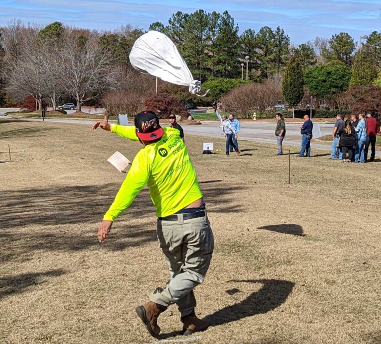 Guy with a backwardsbasebale cap throwing a white sandbag int he air.